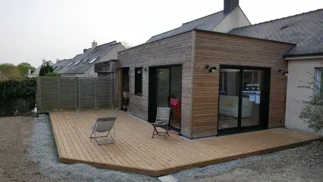 Extension bois et terrasse : transformation moderne de la maison Extension cubique avec bardage bois clair, reliée à une terrasse en bois. Grandes baies vitrées coulissantes noires donnant sur le jardin.